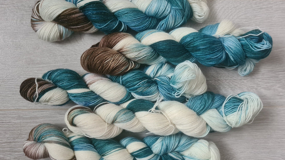 Five skeins of variegated yarn arranged on a light gray wood-textured surface. The yarn features variegated colors of teal, blue, cream, and brown.